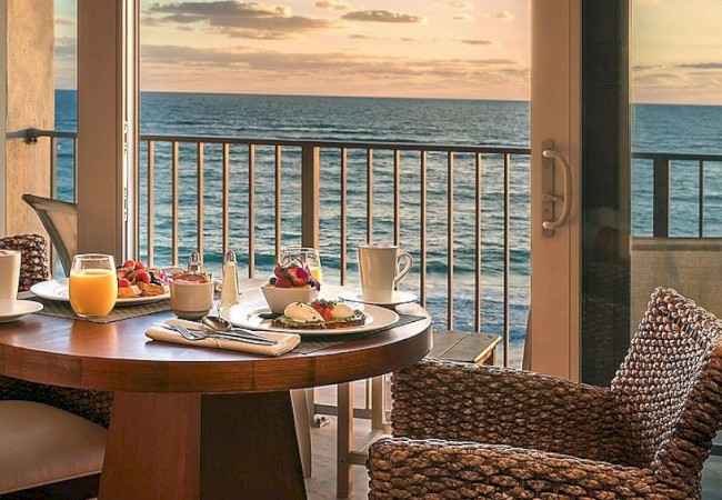 A table set for breakfast with juice, coffee, and fruit, overlooking an ocean view through glass doors during sunset.