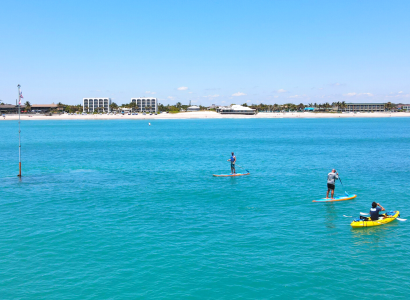 People paddleboarding and kayaking on a calm sea near a sandy beach with buildings in the distance under a clear blue sky.