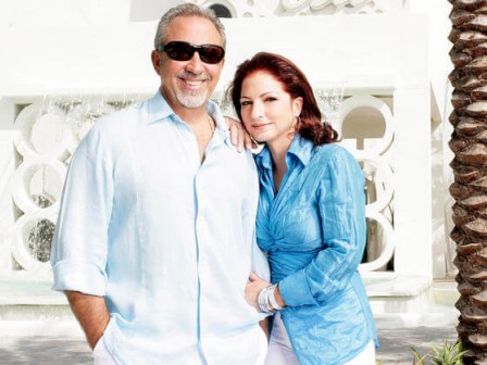 A couple standing in front of a white building with circular designs, palm trees around, both in casual attire smiling at the camera.