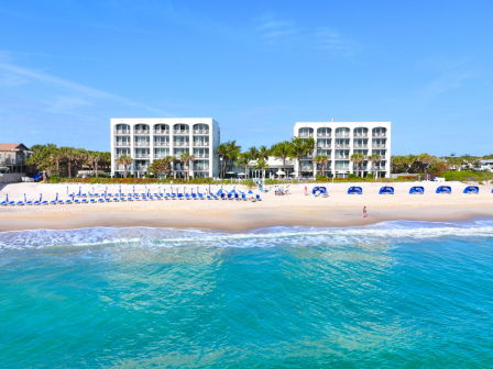 A beachfront scene with two modern white buildings, lined beach chairs, blue umbrellas, palm trees, and clear blue ocean and sky.
