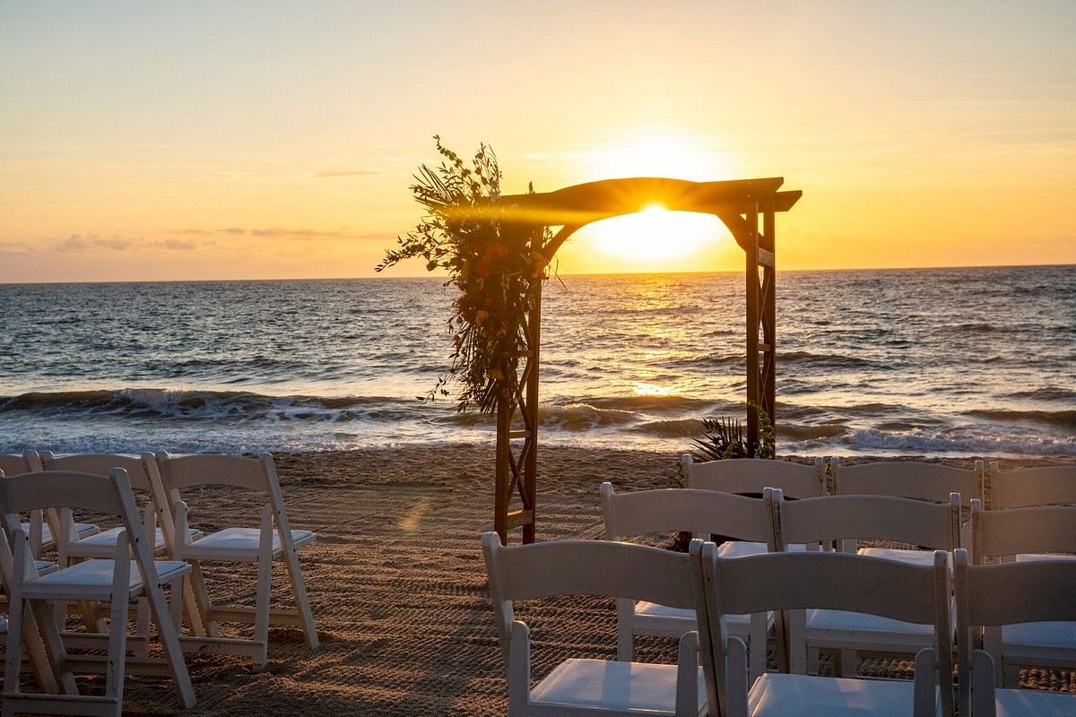 A beach setup for a sunset wedding includes an arch and rows of white chairs facing the ocean.