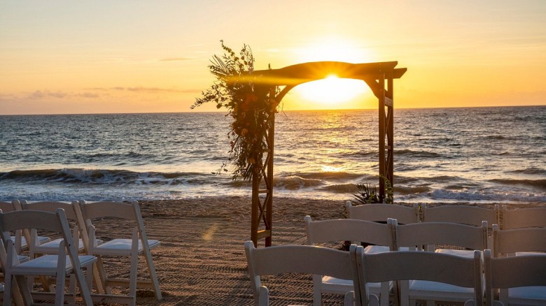 A beach setup for a sunset wedding includes an arch and rows of white chairs facing the ocean.